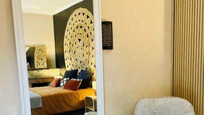 Una habitación con un espejo grande, una cama con cabecera, una silla, un tapete tejido y una lámpara fijada a la pared.