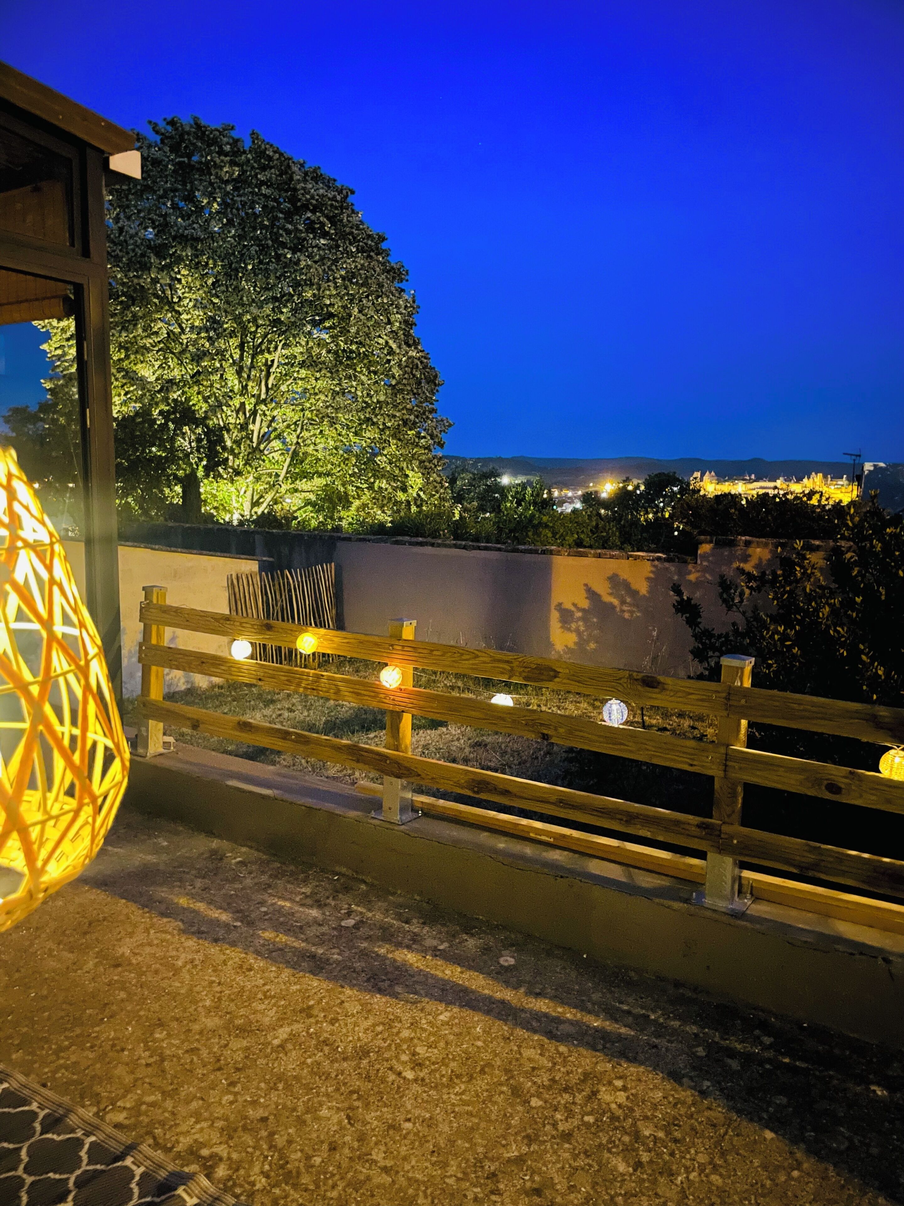 A nighttime view of a coastal area with a wooden fence, a large tree, and distant city lights.