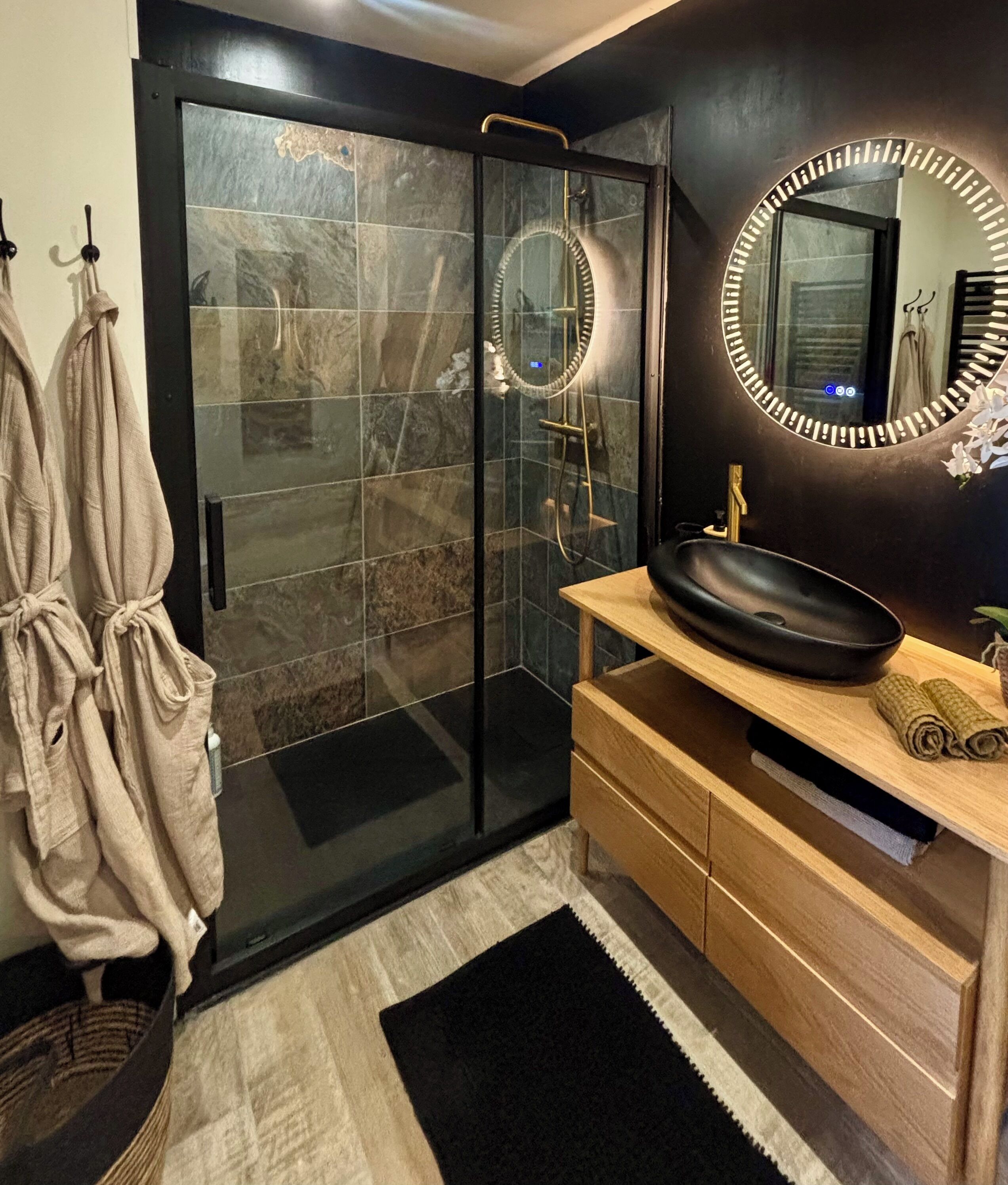 Une salle de bain moderne avec une douche à parois de verre, un grand miroir rond avec éclairage intégré, un lavabo noir, des meubles en bois et un tapis noir.