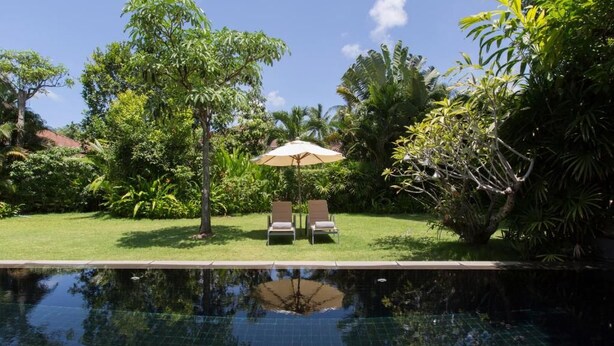 A swimming pool with two lounge chairs and an umbrella, surrounded by lush greenery and trees. - Villa Koru (Surat Thani)
