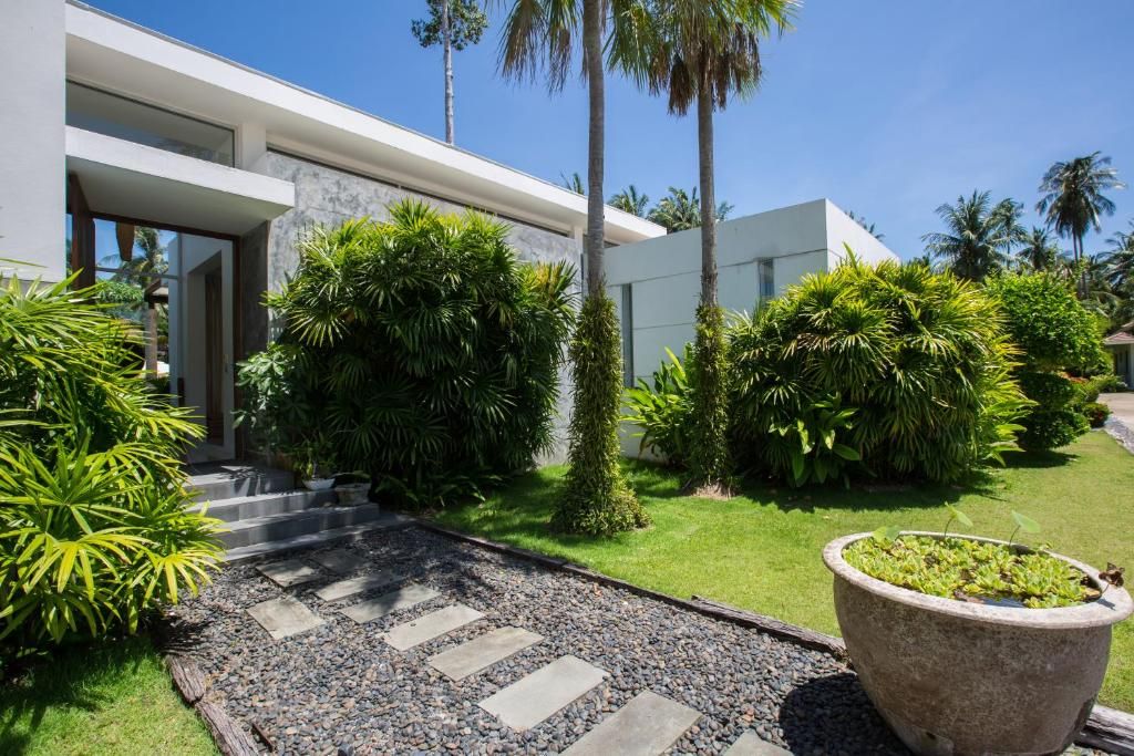 Uma casa moderna com um caminho pavimentado que leva à entrada, rodeada por vegetação exuberante e palmeiras.