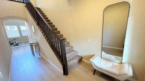 Een trap met een donkeren leuning, een spiegel en een gestoffeerde stoel in een kamer met lichtgekleurde muren en houten vloer.