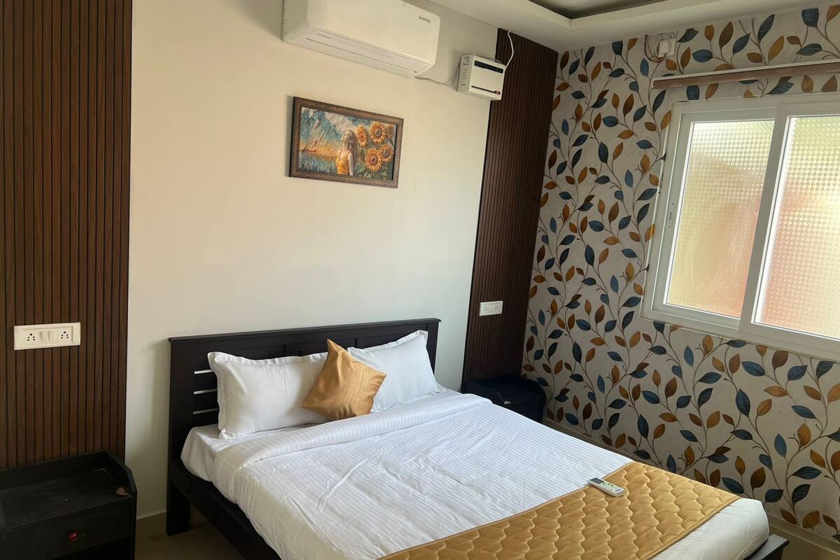 A bedroom with a bed, a nightstand, a wall-mounted air conditioner, a power outlet, a window with a leaf-patterned curtain, and a painting on the wall.