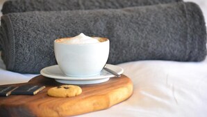 En kopp kaffe på en träbricka, en kaka och en bok på en säng med ett grått täcke och kuddar.