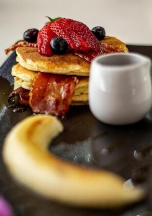 Una pila de tortitas con fresas, arándanos y tocino, acompañada de un vaso de almíbar y una banana.