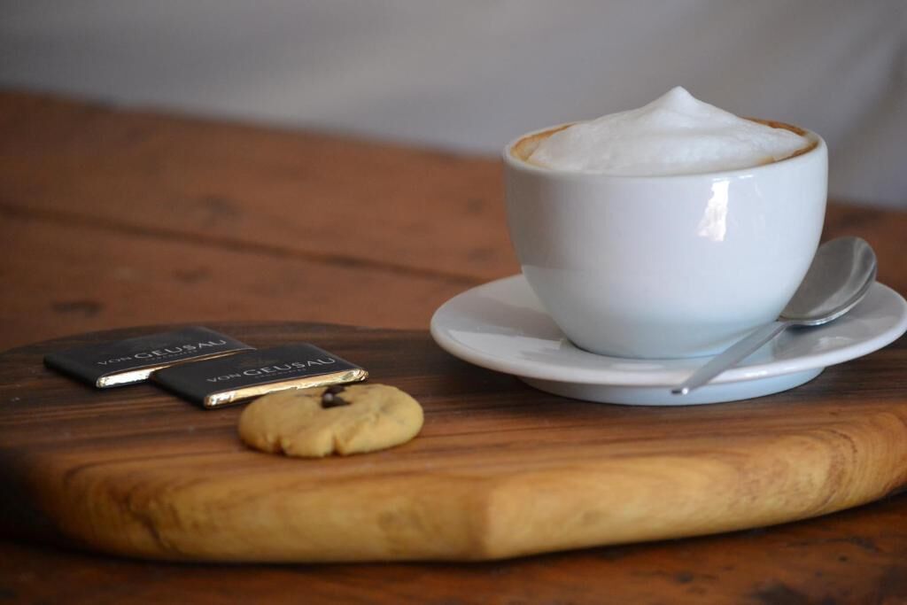 Een witte koffiekop met schuim op een schotel, een lepel, een koekje, en twee kleine zwarte doosjes op een houten tafel.