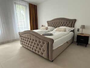 Ein Bett mit geblattetem Kopfteil, ein Nachttisch mit Lampe und ein Fenster mit Vorhängen.