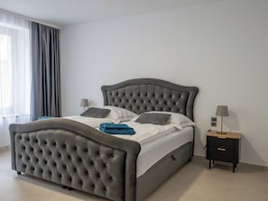 A modern bedroom with a tufted headboard, a bedside table with a lamp, and a view of the outside through sheer curtains. - Ferienwelt Wittenberge - Elbe Apartment (wittenberge)
