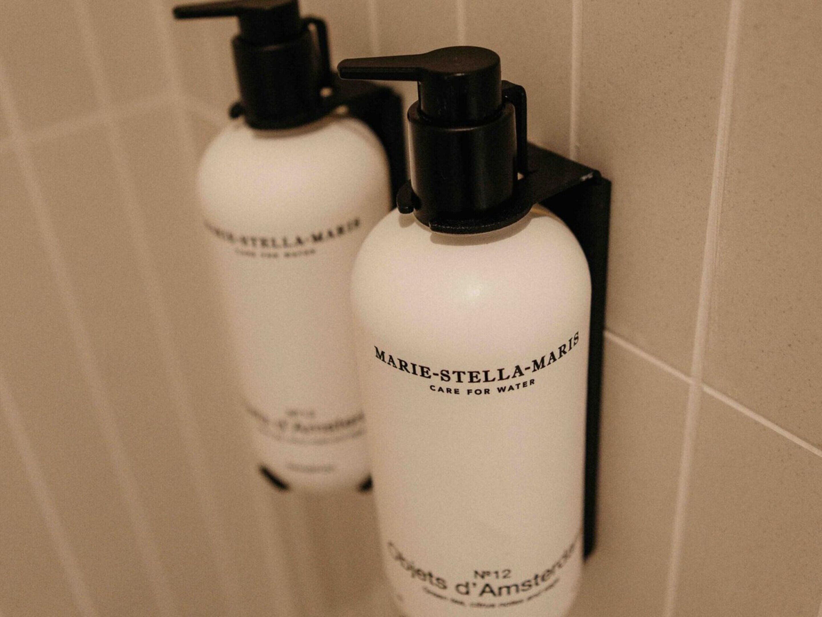 Twee witte flessen met zwarte pompen, voorzien van de aanduiding 'Marie-Stella-Maris', bevestigd aan een betegelde muur.