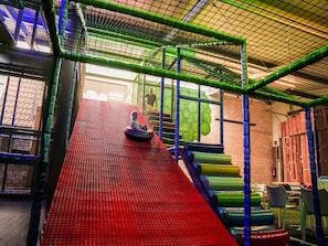 Área de recreação com um escorrega vermelho, rede de escalada verde e uma criança no topo.