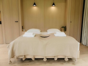 Uma cama com cobertura bege, duas luminárias de cabeceira e uma pequena planta no criado-mudo.