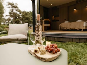 Eine Flasche B.A.C.O.N. Wein, zwei GlÀser, ein Holzbrett mit einer verpackten Sache und Trauben sowie ein Stuhl im Freien.