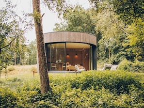 A modern, circular wooden structure with glass walls, surrounded by lush greenery.