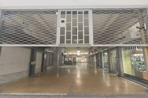 A corridor with a metal security gate, glass storefronts, and a tiled floor. - Modern Studio l Worn Bldg Entrance (Athina)