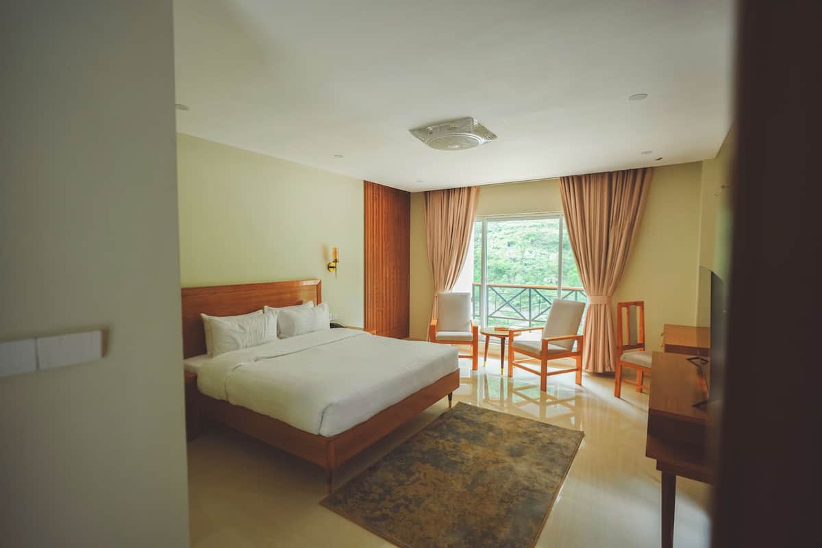 A hotel room with a bed, a desk, a chair, and a view of greenery outside.