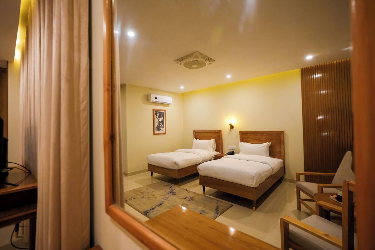 A hotel room with two beds, a desk, a chair, a wall-mounted air conditioner, and a framed picture.