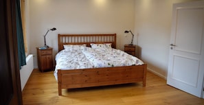 Una cama de madera con una colcha estampada con motivos florales, dos mesitas de noche con lámparas, una puerta blanca y un suelo de madera.
