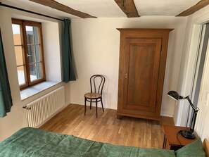 A room with a wooden floor, a radiator, a window with curtains, a wooden wardrobe, a chair, a desk with a lamp, and a bed with a green bedspread. - Quiet and central in a historic house with a variety of excursion destinations (Kandern)