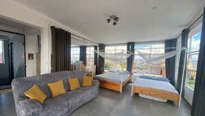 A modern hotel room with a grey sofa, two single beds, a large window with a city view, and a ceiling fan. - Apartment with 360-degree view (Antananarivo)
