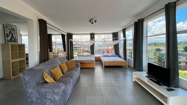 A modern living room with a grey sofa, a wooden bed, a TV on a stand, and large windows offering a city view. - Apartment with 360-degree view (Antananarivo)