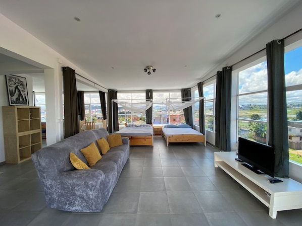 A modern living room with a grey sofa, a wooden bed, a TV on a stand, and large windows offering a city view.
