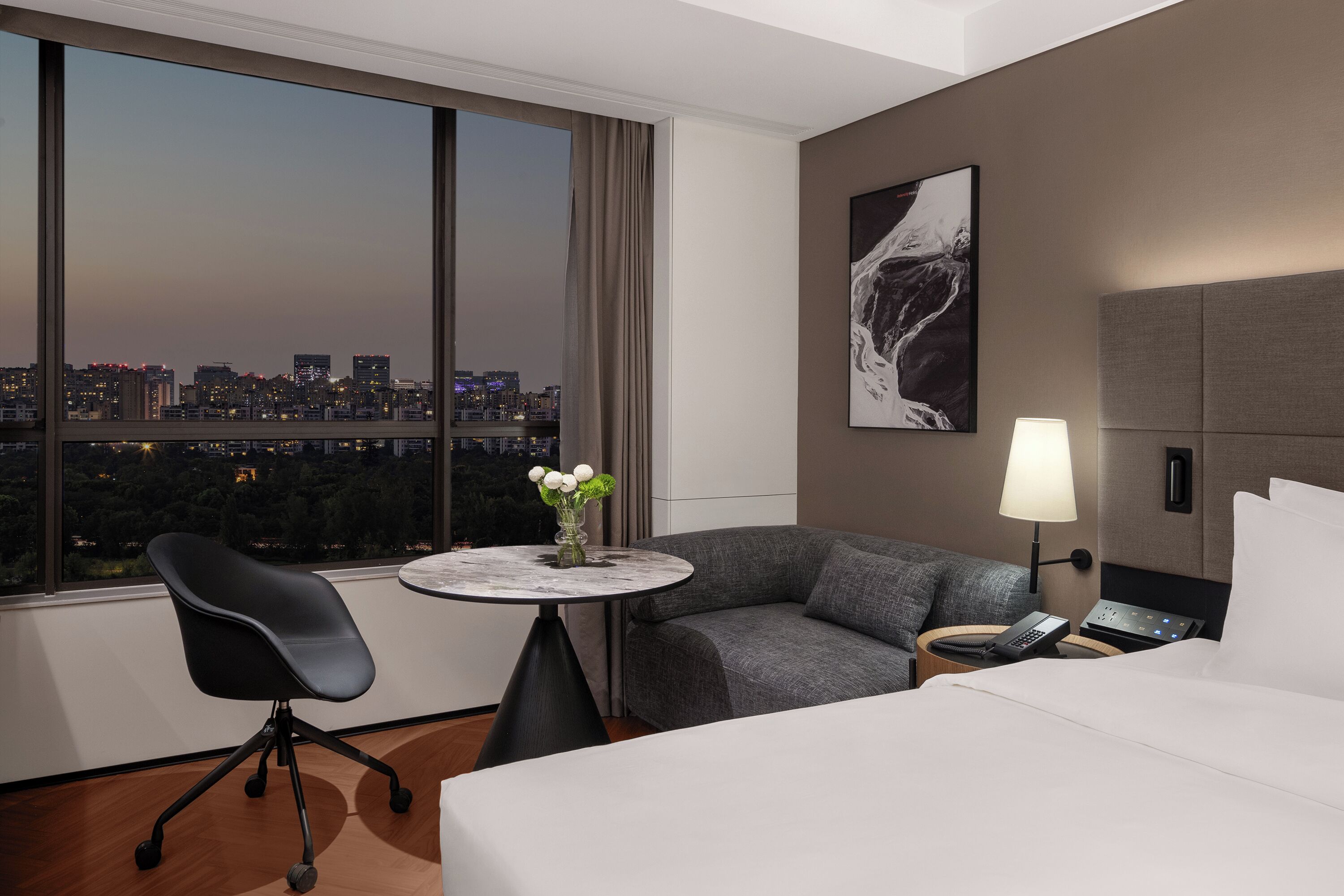 Habitación de hotel con cama, escritorio, silla, mesa redonda con un florero, sofá, lámpara y un cuadro en la pared.