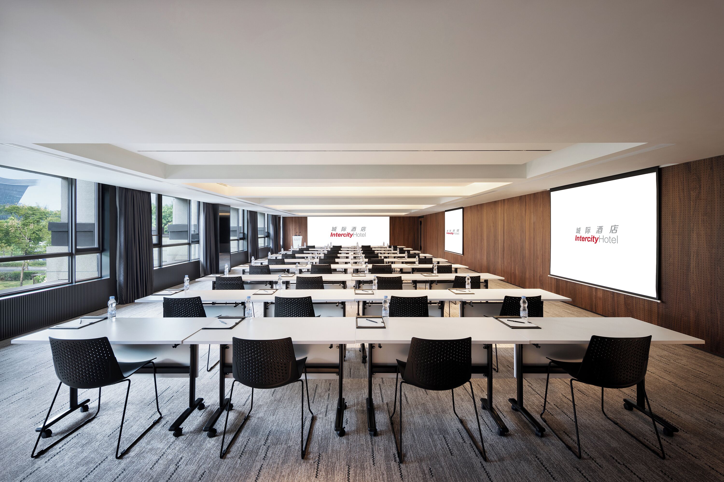 Un salón de conferencias con varias mesas y sillas, ventanas amplias y una pantalla de proyección que muestra 'Intercity Hotel'.