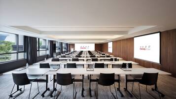 Un salón de conferencias con varias mesas y sillas, ventanas amplias y una pantalla de proyección que muestra 'Intercity Hotel'.