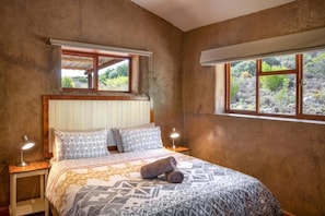 A bedroom with a bed, two bedside lamps, a window with a view, and a wooden headboard. - Kingfisher Cottages (Montagu)