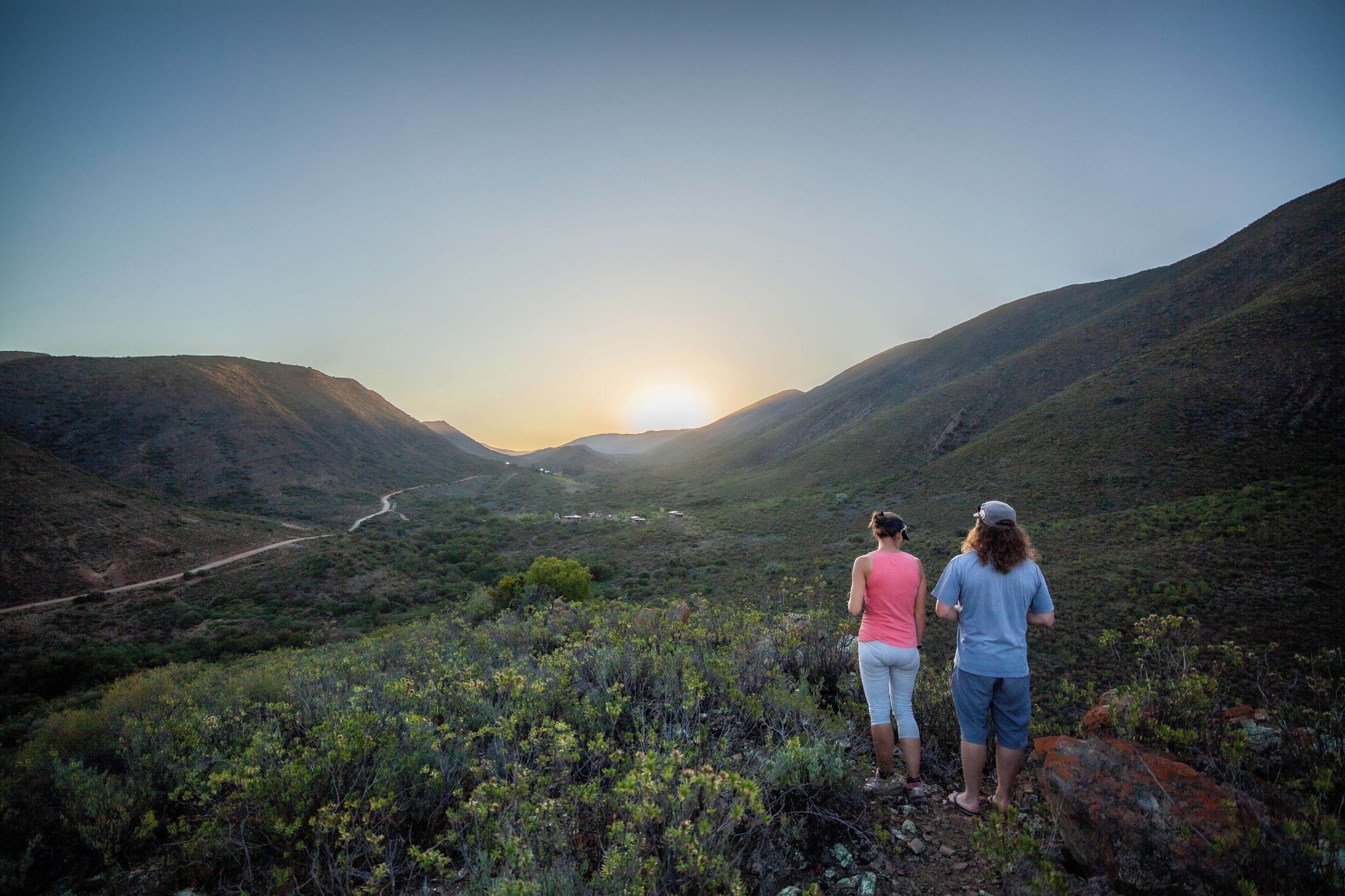 Two people standing on a hill overlooking a valley with a road leading into the distance and the sun setting in the background.
