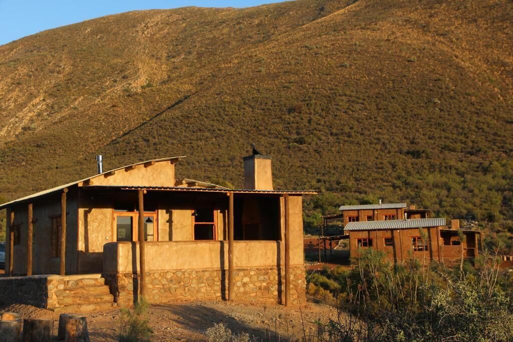 A rustic two-story building with a stone foundation and a flat roof, surrounded by a dry, hilly landscape.