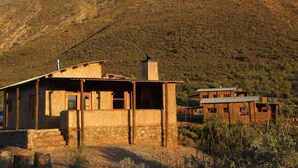 A rustic two-story building with a stone foundation and a flat roof, surrounded by a dry, hilly landscape.