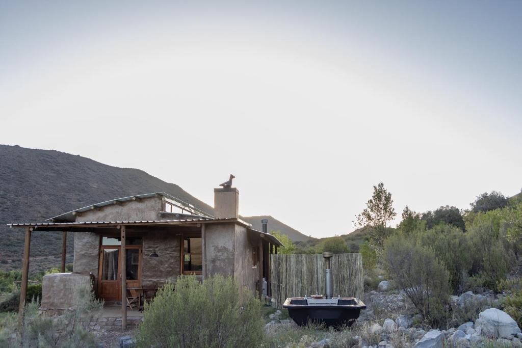 Una cabaña rústica con porche de madera, chimenea y un jacuzzi en un entorno natural.