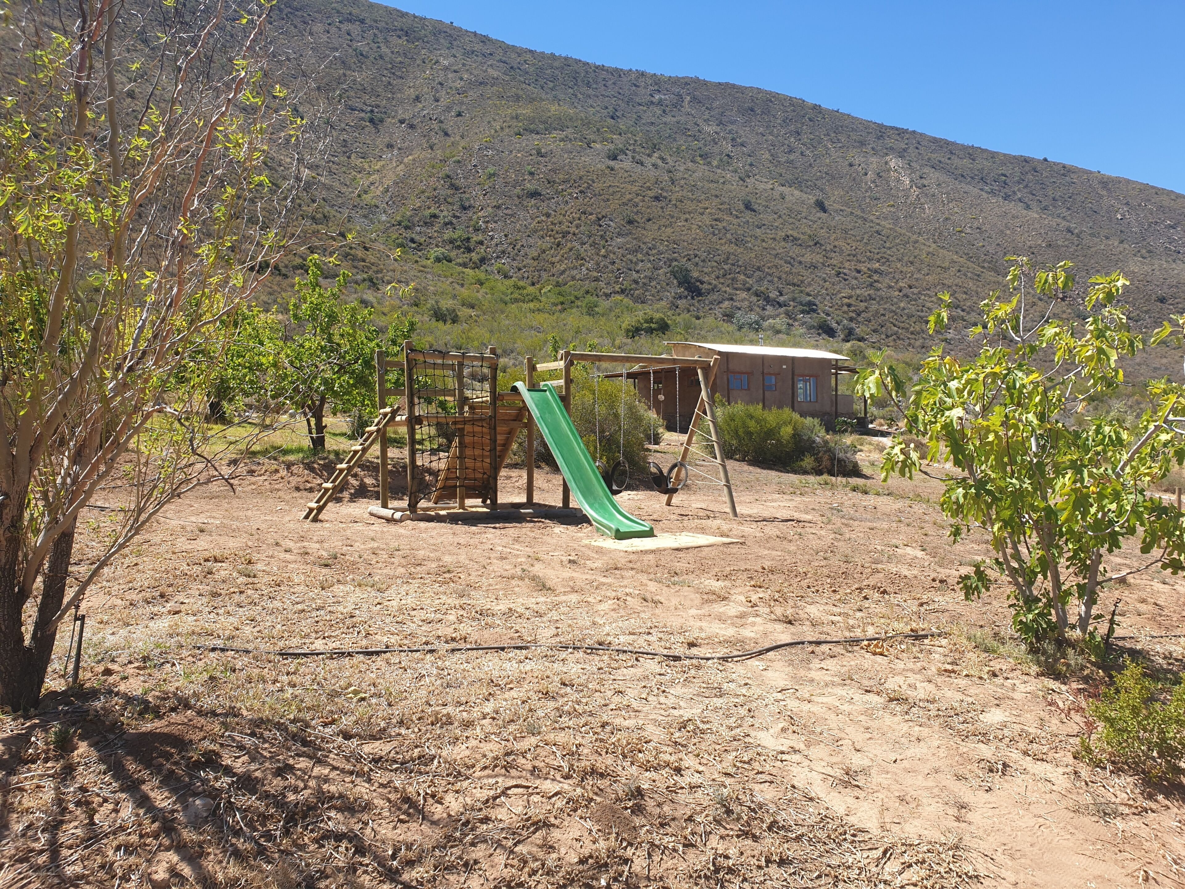 A playground with a slide and climbing structures, surrounded by dry grass and sparse trees, with a hill in the background.