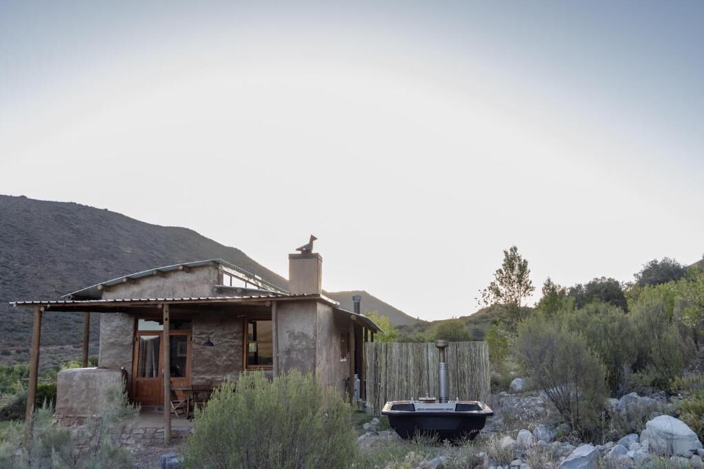 Une cabane rustique avec une véranda en bois, une cheminée et un réservoir d’eau, située dans un paysage désertique.