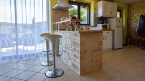 A kitchen with a central island, white cabinets, a microwave, a coffee maker, and a refrigerator. There is a bar stool and a window with a view of the outside. - Holiday Home in Montcléra with Barbecue (Montclera)