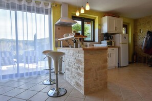 Eine Küche mit einer Kochinsel, weißen Schränken, einer Mikrowelle, einer Kaffeemaschine und einem Kühlschrank. Ein Barhocker und ein Fenster mit Blick ins Freie sind ebenfalls vorhanden.