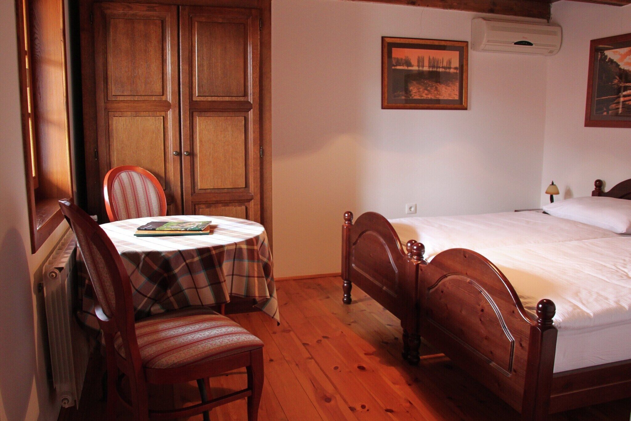 Ein Schlafzimmer mit einem hölzernen Bett, einem Sessel, einem Tisch mit karierter Tischdecke und einem Kleiderschrank.
