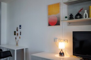 Un salon moderne avec un bureau blanc, une lampe, un téléviseur à écran plat et des objets décoratifs disposés sur des étagères.