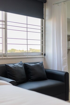 Una habitación con una cama, un sofá negro con dos cojines, una ventana con cortina blanca y vistas al exterior.