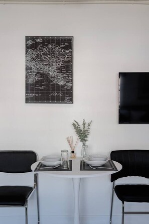 Un comedor con una mesa redonda, dos sillas negras, un mapa mundial, un florero con plantas y un televisor montado en la pared.