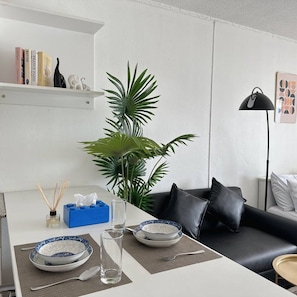 Ein modernes Wohnzimmer mit Essbereich, einer schwarzen Ledersofa, einem Couchtisch, einem Bücherregal und einer großen Grünpflanze.