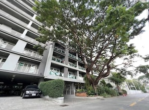 A multi-story building with balconies, a parking garage, and a large tree in front. - Stay Classy – Pool• Gym• Balcony (Chiang Mai)