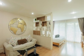 A modern living room with a sofa, a glass coffee table, a world map wall art, a built-in bookshelf, a bed with a canopy, and a large window with curtains. - Stay Classy – Pool• Gym• Balcony (Chiang Mai)