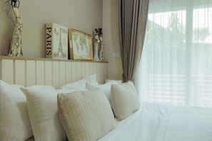 A neatly made bed with white linens and several pillows, a shelf with decorative items and framed pictures, and a window with curtains. - Stay Classy – Pool• Gym• Balcony (Chiang Mai)