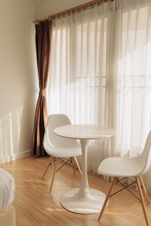 Ein Zimmer mit einem runden Tisch, zwei Stühlen, einem Fenster mit durchsichtigen Vorhängen und einem Holzboden.