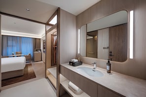 A modern hotel room with a large bed, a bathroom with a large mirror, and a desk with a chair.