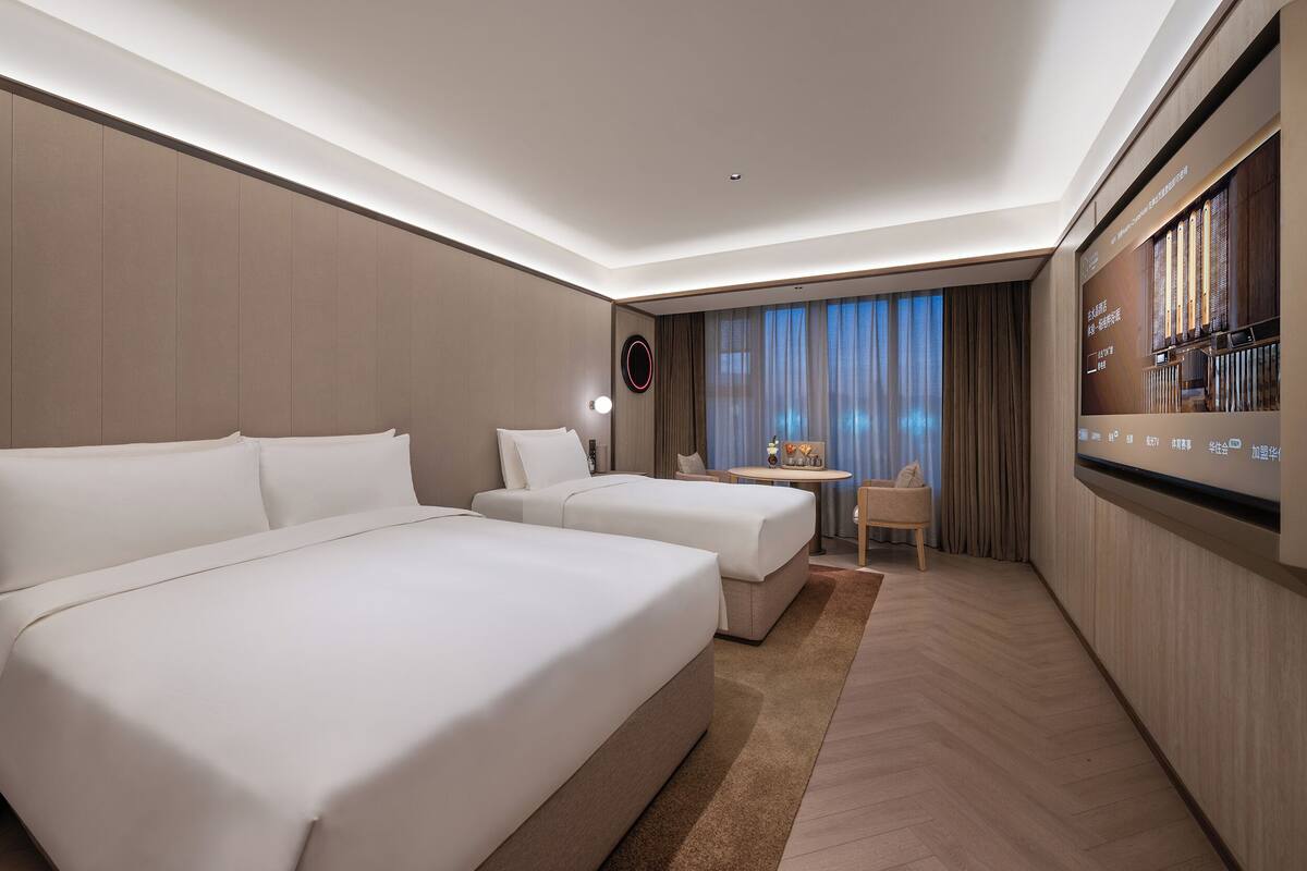 A hotel room with two beds, a small table, a chair, and a television mounted on the wall.