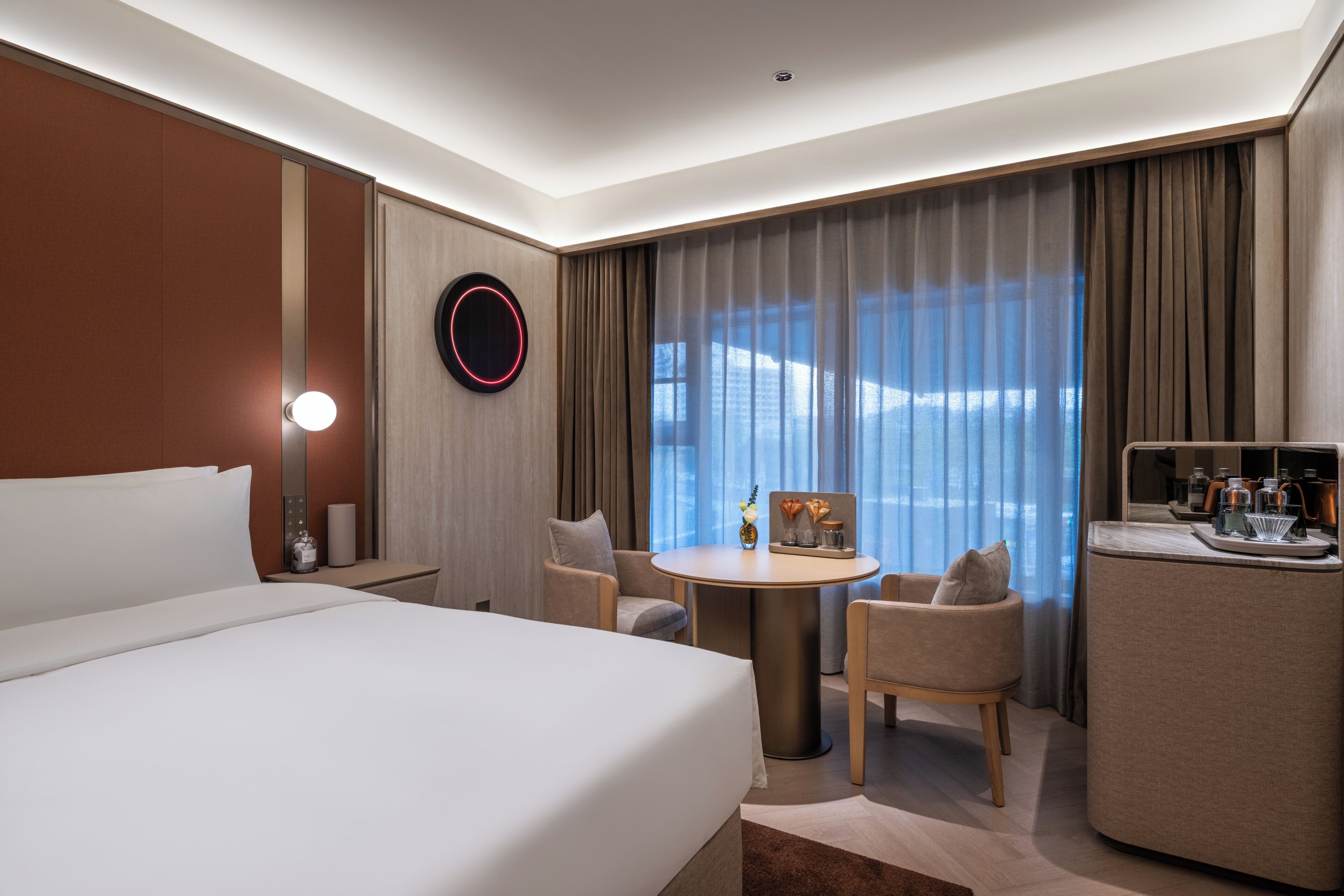 A hotel room with a bed, a round table, two chairs, a small table with a vase, a mirror, and a window with curtains.