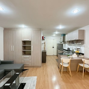 A modern kitchen with wooden cabinets, a dining area with chairs, and a sofa in a living room. - Spacious Studio with Balcony in Nimman Soi 15 | Pool, Gym & Washer (Chiang Mai)